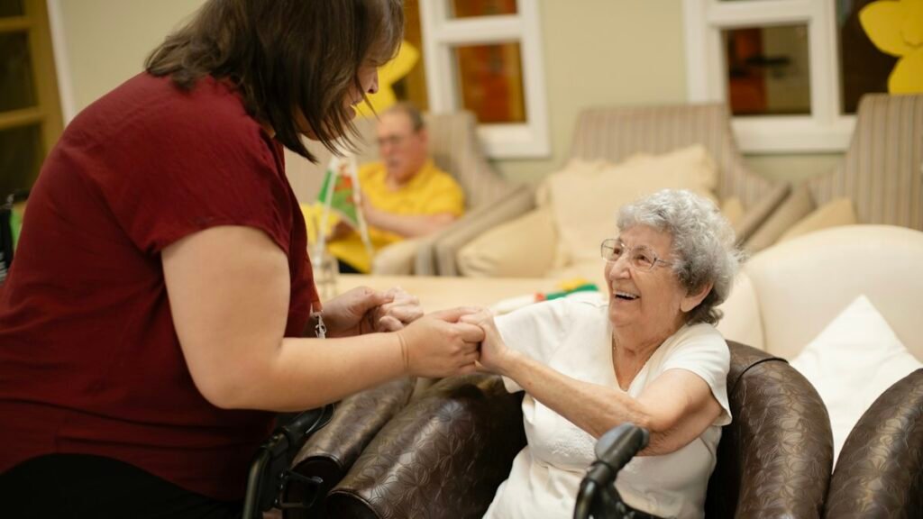 A caregiver holds the hands of a smiling older woman seated with her walker at home