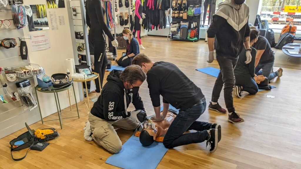 Pairs of trainees practise CPR on manikins during a group first aid class, with an AED training kit on the floor